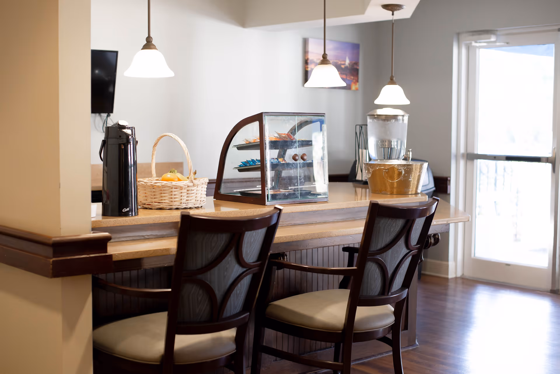 A communal serving counter with two chairs, pendant lights, a glass pastry case, beverage dispenser and a fruit basket in a dining area.