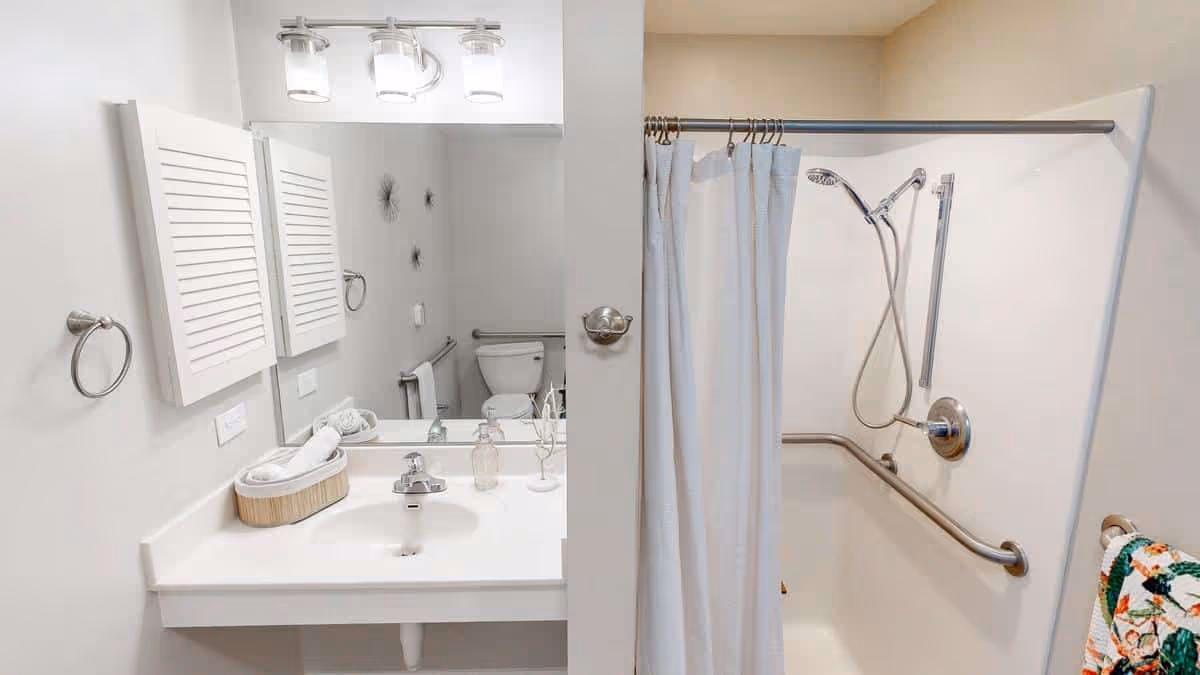 A clean and modern bathroom featuring a white sink with a silver faucet, a large mirror above the sink, and a light fixture with three lights. To the right, there is a bathtub with a white shower curtain, a handheld showerhead, and safety grab bars. A toilet is visible in the background, along with a towel rack holding a colorful towel.