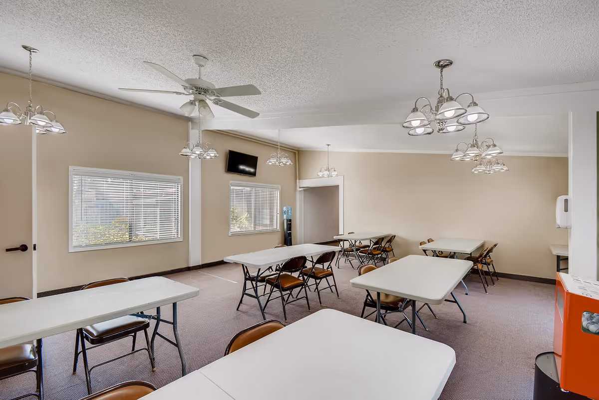 Communal dining/activity room with folding tables and chairs, chandeliers, windows, and a wall-mounted TV.