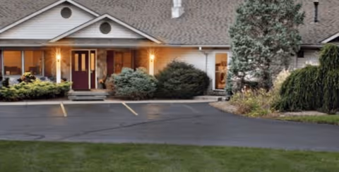 Front entrance of a single-story senior living building with lit porch lights, shrubs, and a small parking area.