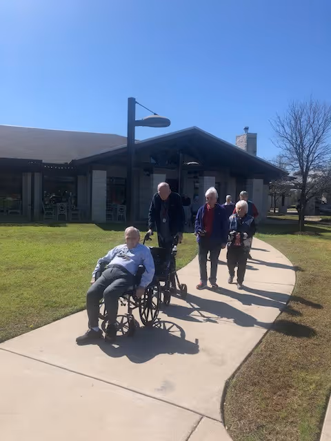 A group of elderly people walking on a curved sidewalk outside a building on a sunny day. One person is seated in a wheelchair being pushed by another. The building has a stone exterior with a covered entrance and outdoor seating visible inside.