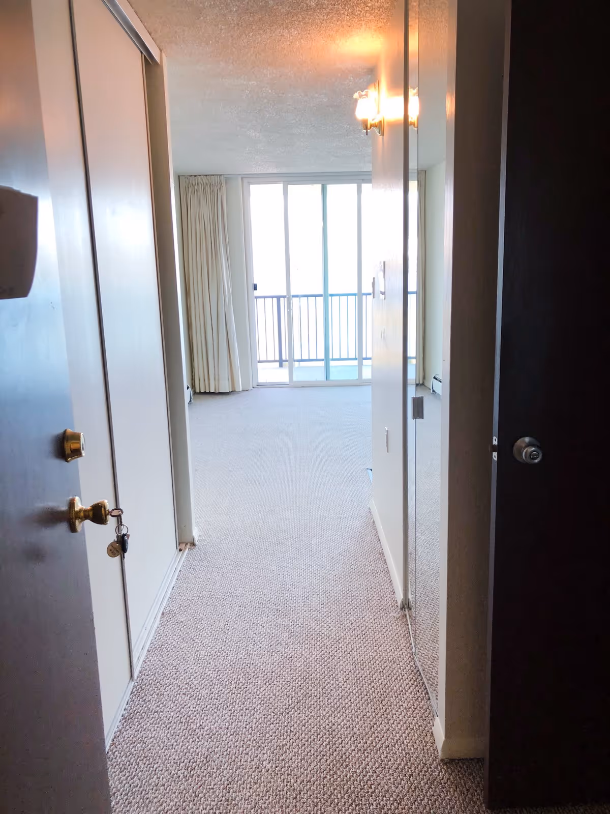 View down a carpeted hallway in a senior living facility with an open door on the left showing a closet with a key in the lock, a wall-mounted light fixture on the right, and large sliding glass doors at the end leading to a balcony with curtains on either side.