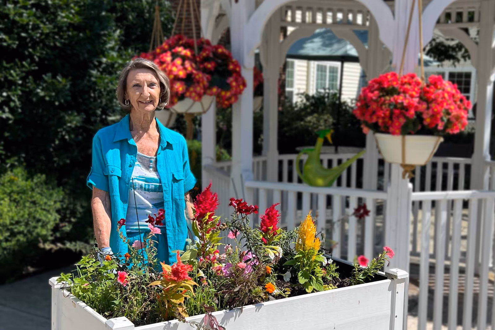 An elderly woman wearing a turquoise shirt and gloves stands behind a white planter box filled with colorful flowers. She is outdoors near a white gazebo adorned with hanging flower baskets and a green watering can. The background includes greenery and part of a building.