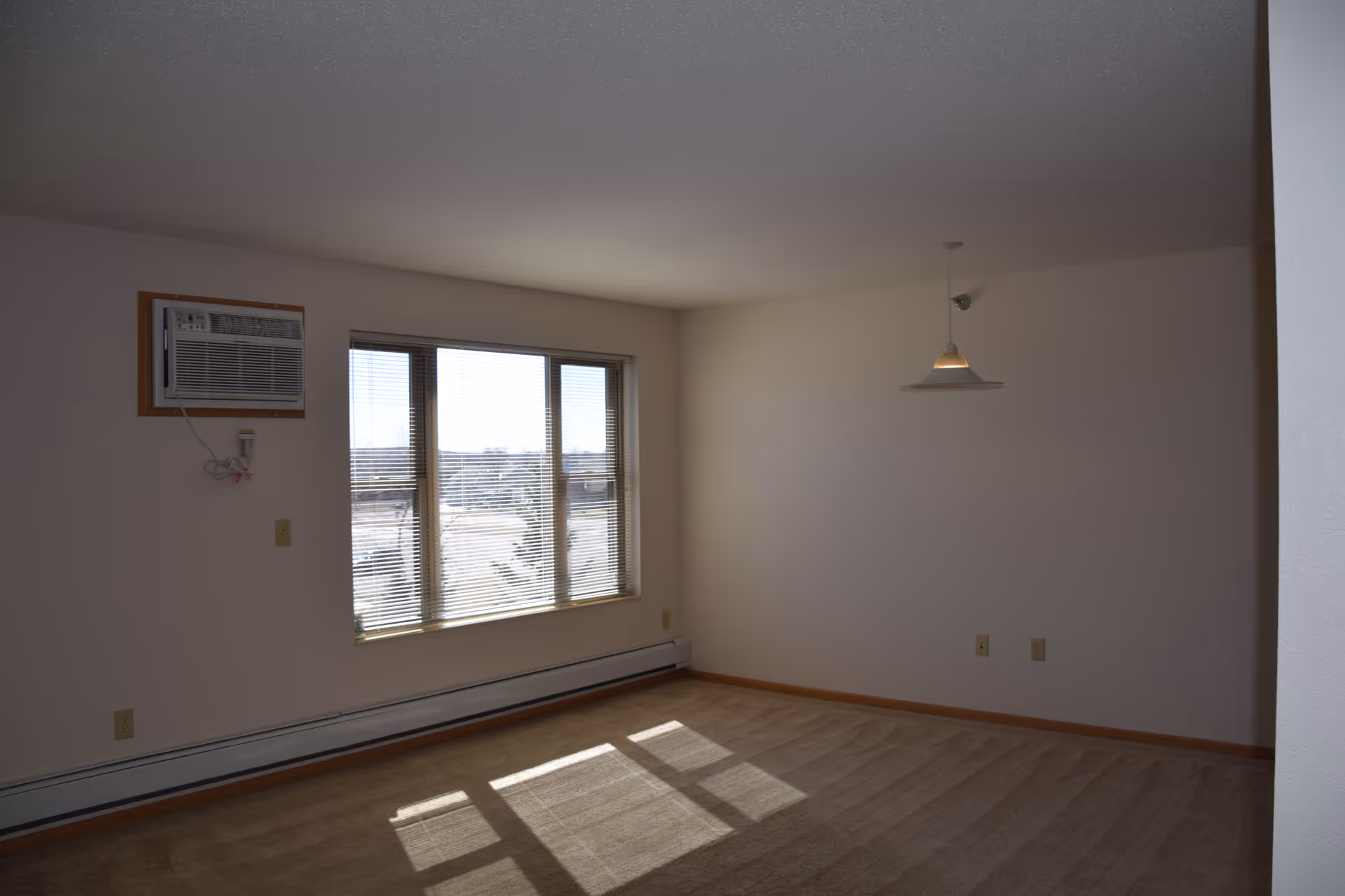 Empty room with beige carpet, white walls, a window air conditioning unit mounted on the wall, and a large window with blinds letting in sunlight. A hanging ceiling light fixture is visible on the right side of the room.