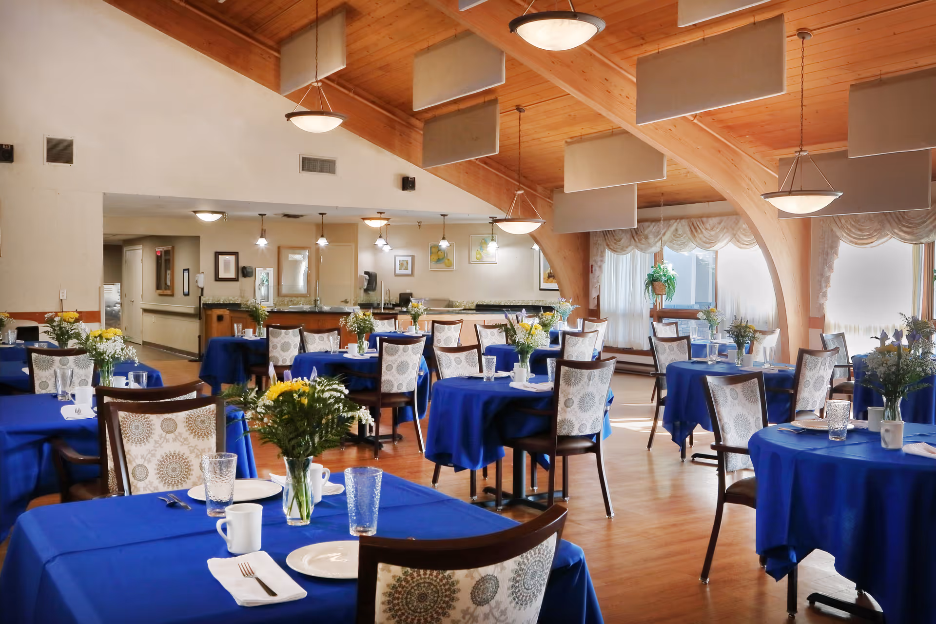 A bright dining room with several tables covered in blue tablecloths, each set with plates, glasses, and floral centerpieces. The room features wooden floors, a high wooden ceiling with hanging light fixtures, and large windows with sheer curtains allowing natural light to fill the space.