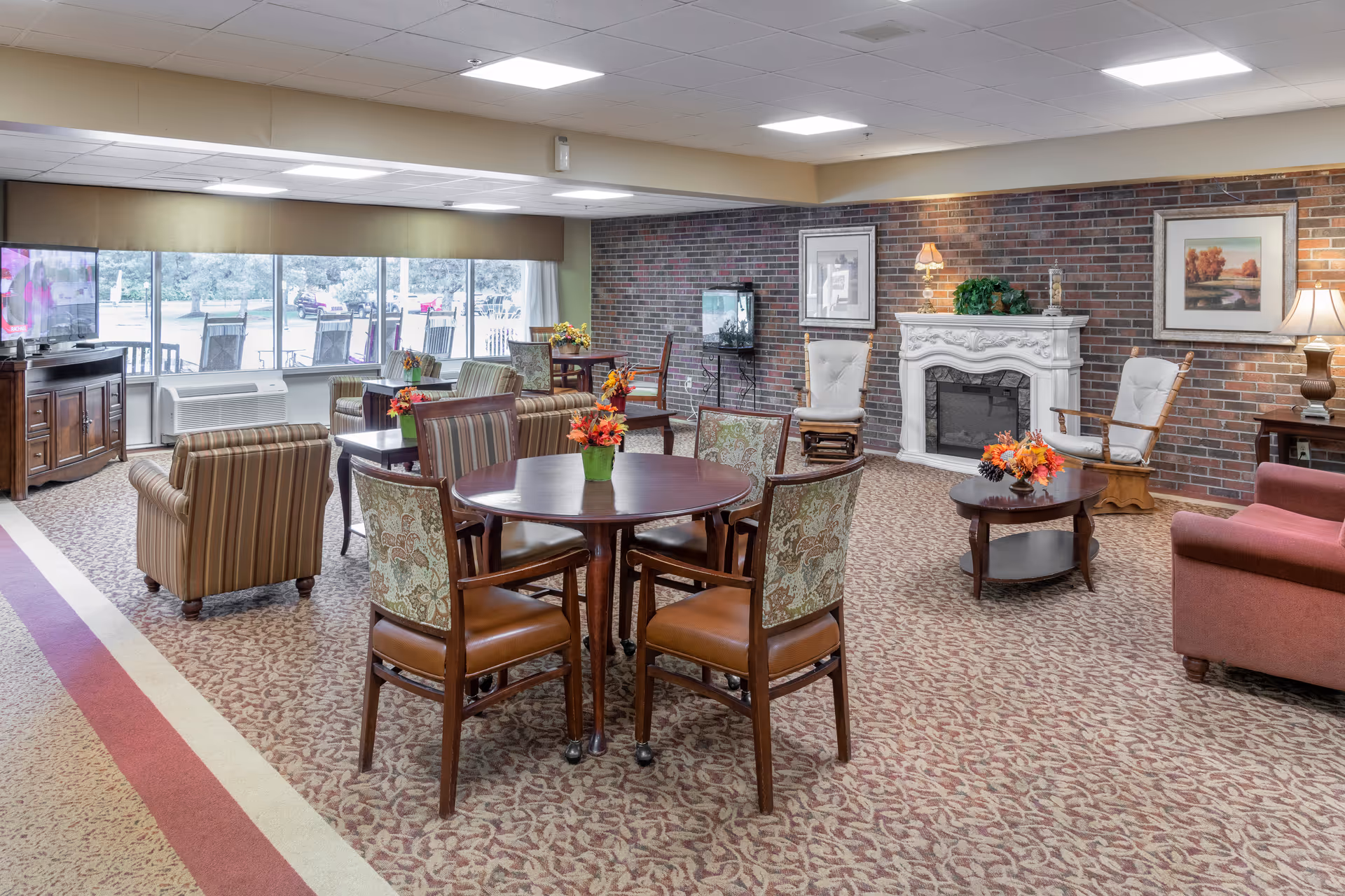 A spacious senior living common area with patterned carpet, multiple round tables with floral centerpieces, upholstered chairs, armchairs, a white decorative fireplace, framed artwork on a brick wall, and large windows overlooking an outdoor area.
