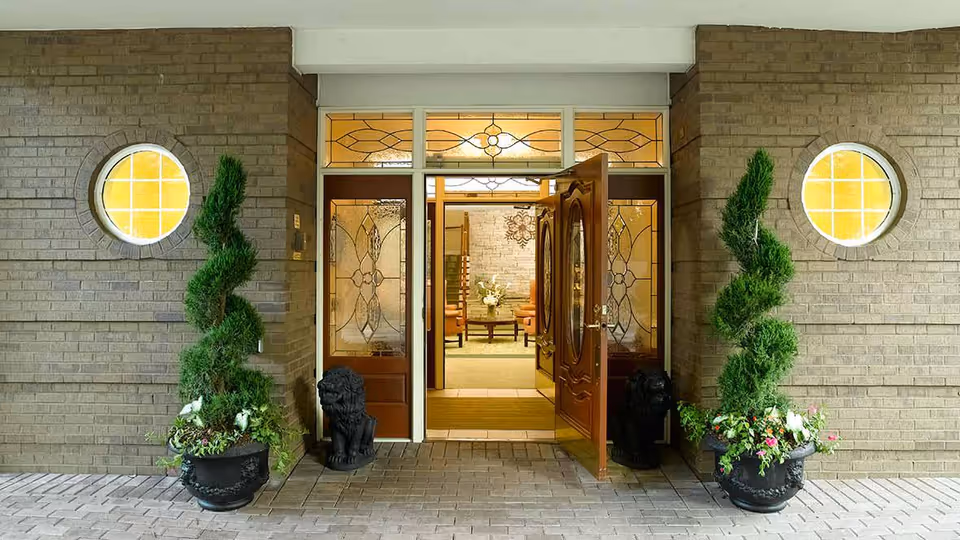 Entrance to a building with double wooden doors, each featuring decorative glass panels. Two spiral-shaped topiary plants in large black pots flank the doorway, with two black lion statues beside the plants. The exterior walls are made of light brown bricks with round windows on either side of the door. Inside, a warmly lit room with chairs and a table with a flower arrangement is visible.