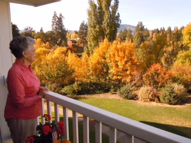 An elderly woman wearing a pink jacket stands on a balcony overlooking a garden with green grass and trees with autumn foliage in shades of yellow, orange, and red. The scene includes a clear sky and distant hills.