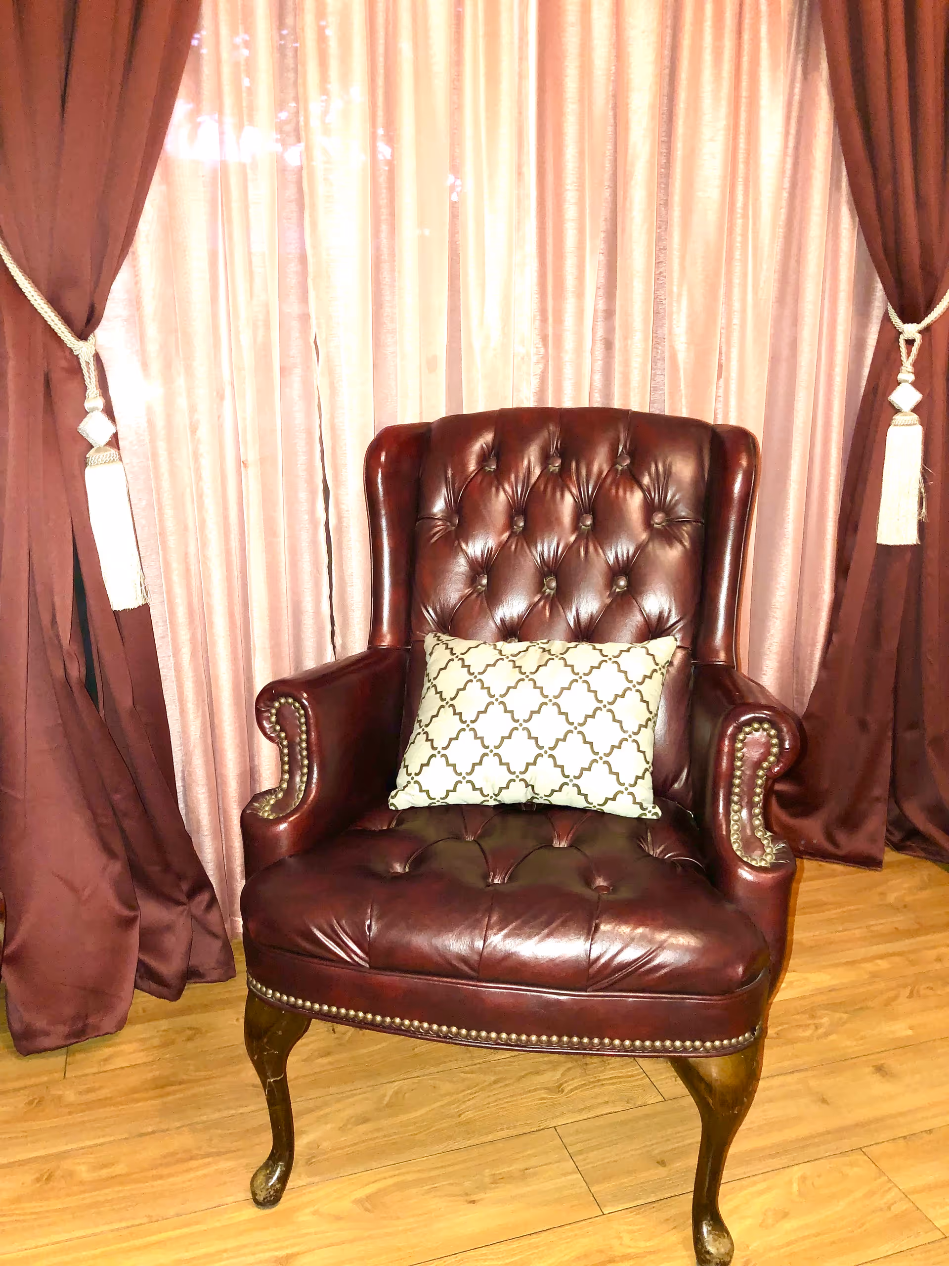 A dark red leather tufted armchair with wooden legs and brass nailhead trim, featuring a decorative white pillow with a black geometric pattern. The chair is placed on a wooden floor in front of pink and maroon curtains tied back with white tassels.
