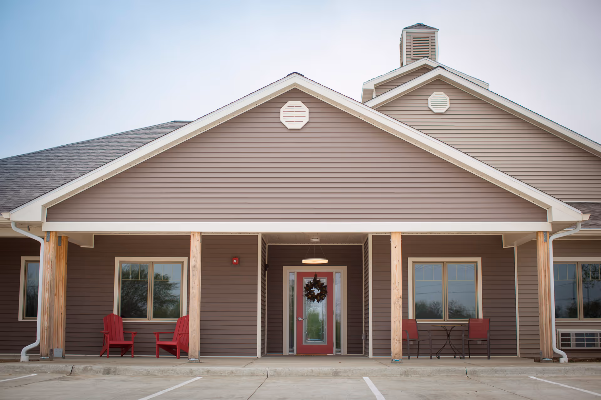 Front exterior view of a single-story building with brown siding and a peaked roof. The entrance features a red door with a wreath, flanked by windows and wooden pillars supporting a covered porch. There are red chairs and a small table on the porch, and a parking area in front.
