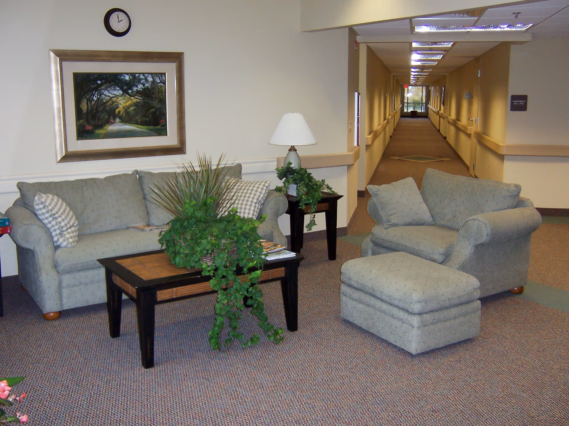 A cozy sitting area in a senior living facility with a light green sofa, matching armchair with ottoman, and a wooden coffee table with a green leafy plant. A side table with a lamp and another plant is next to the sofa. A framed painting of a tree-lined path and a clock are on the wall behind the sofa. A long hallway extends in the background with handrails on both sides and a window at the end.