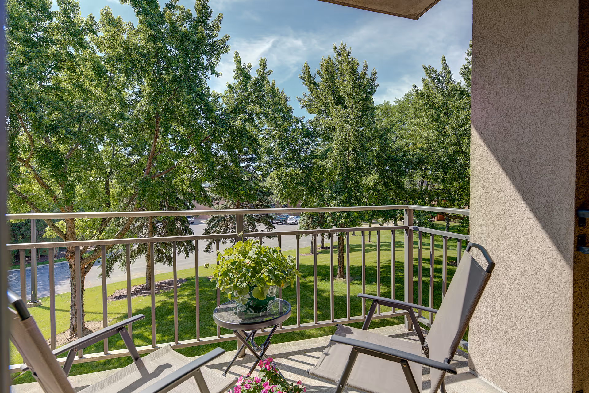 View from a balcony with two chairs and a small round table holding a potted plant, overlooking a green lawn with several trees and a parking lot in the background under a partly cloudy sky.