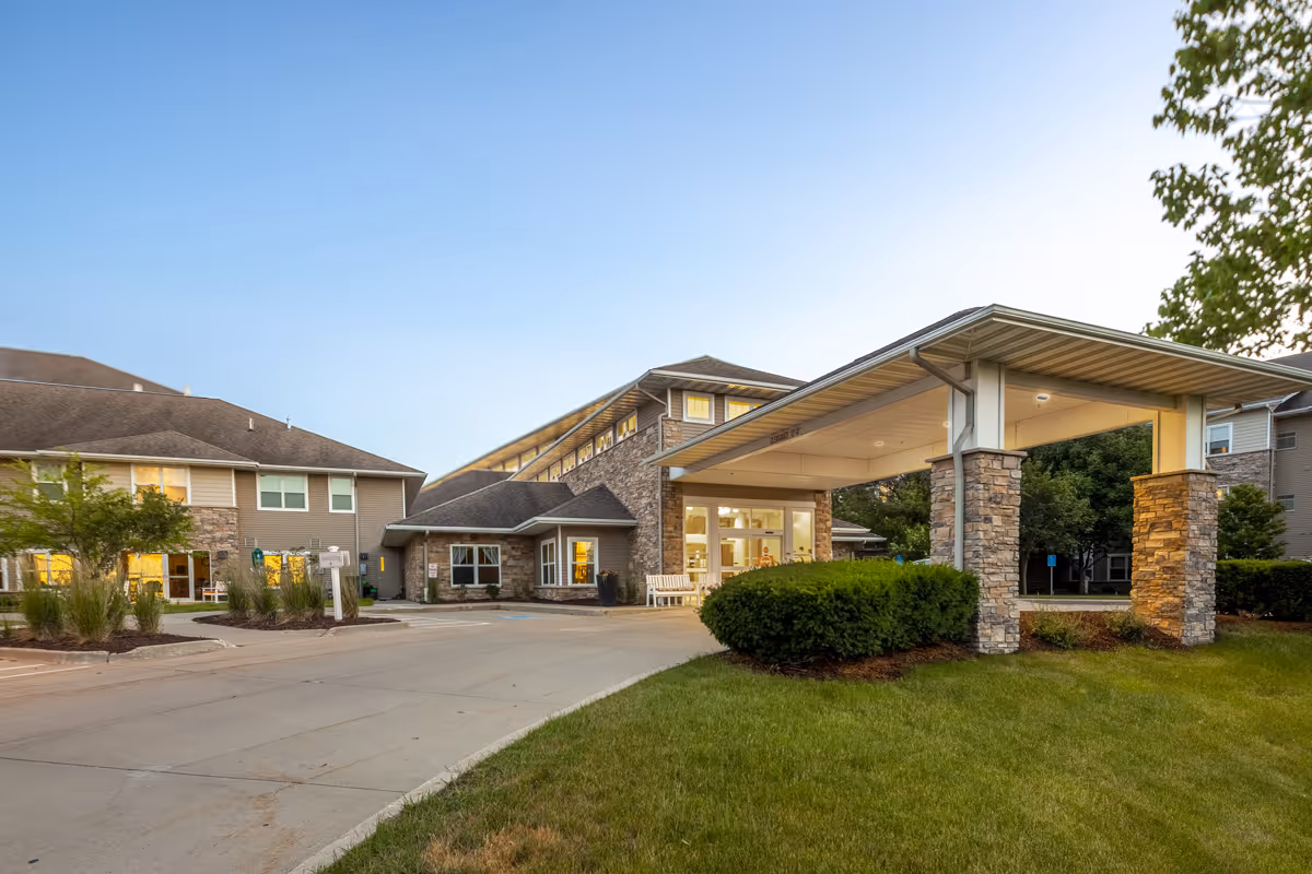 Exterior view of Independence Village of Waukee senior living facility during early evening with a covered entrance supported by stone pillars, well-maintained landscaping, and a clear sky.