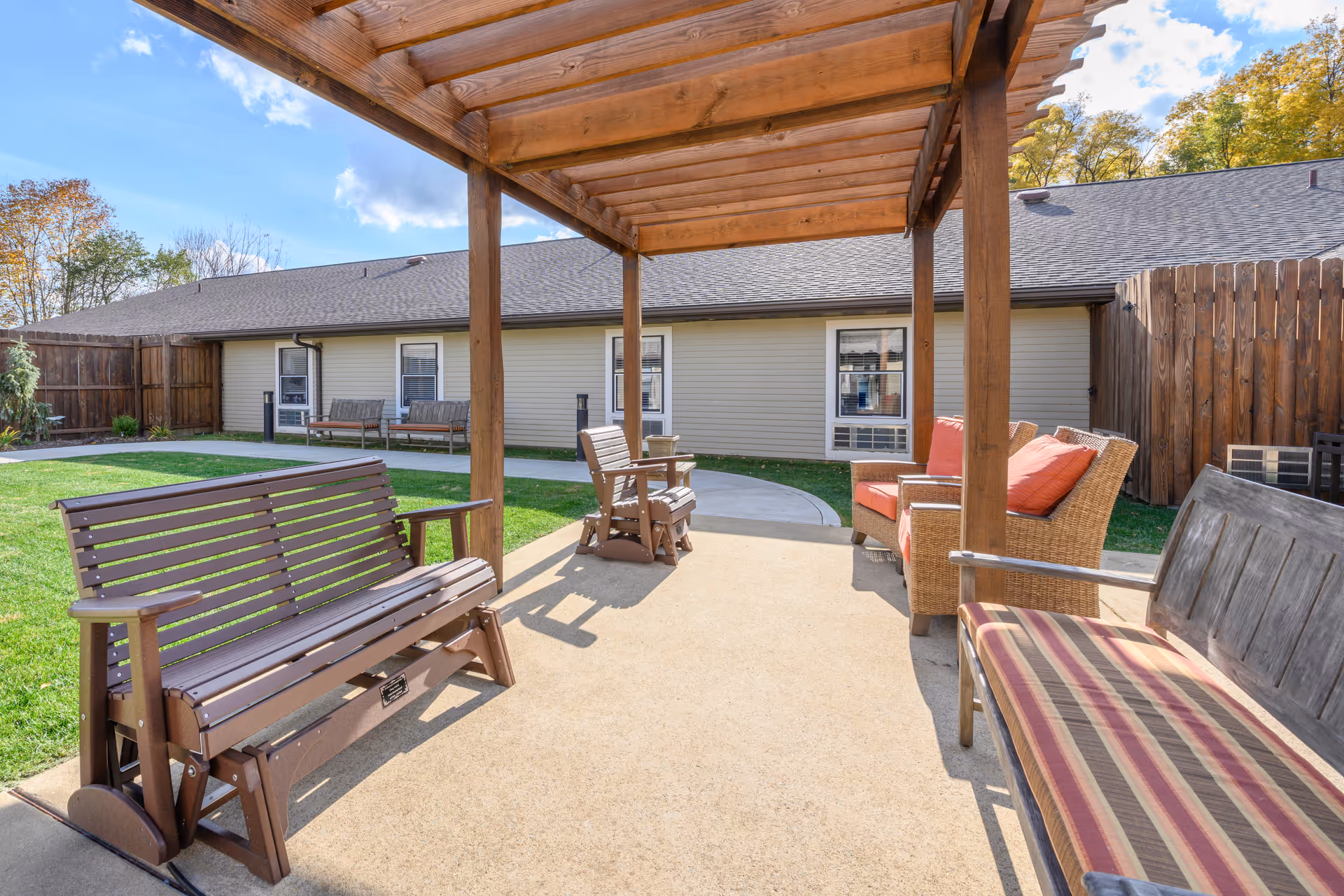 Outdoor seating area at Brookdale Latrobe featuring a wooden pergola, several benches including a brown wooden bench, a striped cushioned bench, and wicker chairs with orange cushions. The area is paved and surrounded by green grass, with a building and wooden fence in the background under a partly cloudy sky.