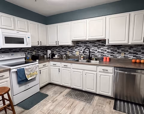 A modern kitchen with white cabinets, a white microwave above a white stove, a stainless steel dishwasher, and a double sink with a black faucet. The backsplash features a mosaic tile design in shades of gray, black, and white. The floor is wood-patterned, and there are various kitchen items on the countertops including a coffee maker, cups, and some fruit.