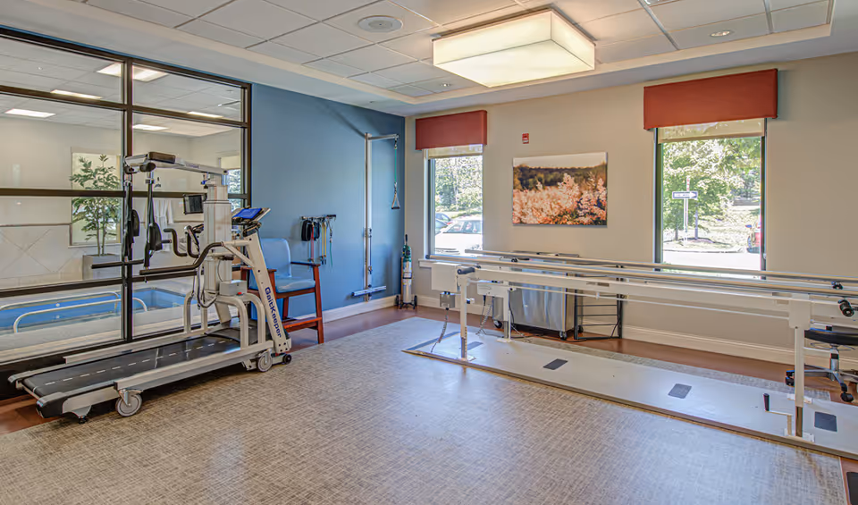 A rehabilitation room with a treadmill and parallel bars for physical therapy. The room has large windows with red valances, a blue accent wall, and a framed picture of flowers. There is also a chair and some medical equipment along the walls.