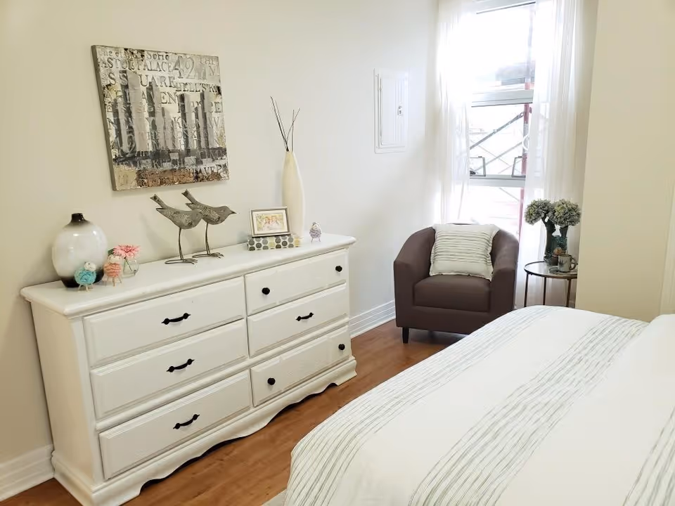 A bright bedroom with a white dresser adorned with decorative items including a vase, bird sculptures, and a framed picture. There is a brown armchair with a striped pillow next to a window with sheer white curtains, and a small round side table holding a vase with flowers. The bed has a white and gray striped comforter.