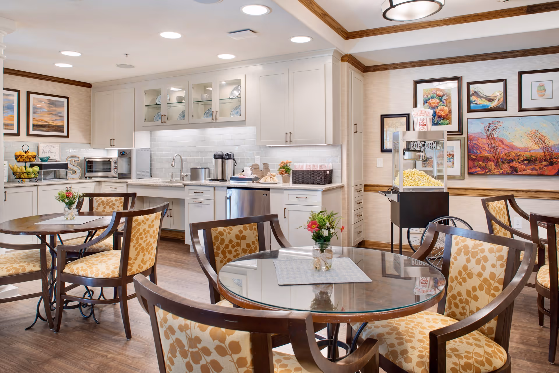 A bright and clean dining area with round glass-top tables and cushioned chairs featuring a yellow leaf pattern. The background shows a white kitchen with cabinets, a microwave, a coffee station, and a popcorn machine. Colorful framed artwork decorates the walls.