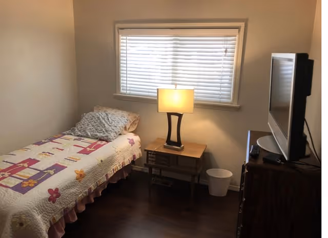 Small bedroom with a twin bed covered in a floral quilt, a nightstand with a lamp beneath a window with blinds, and a TV on a dresser.