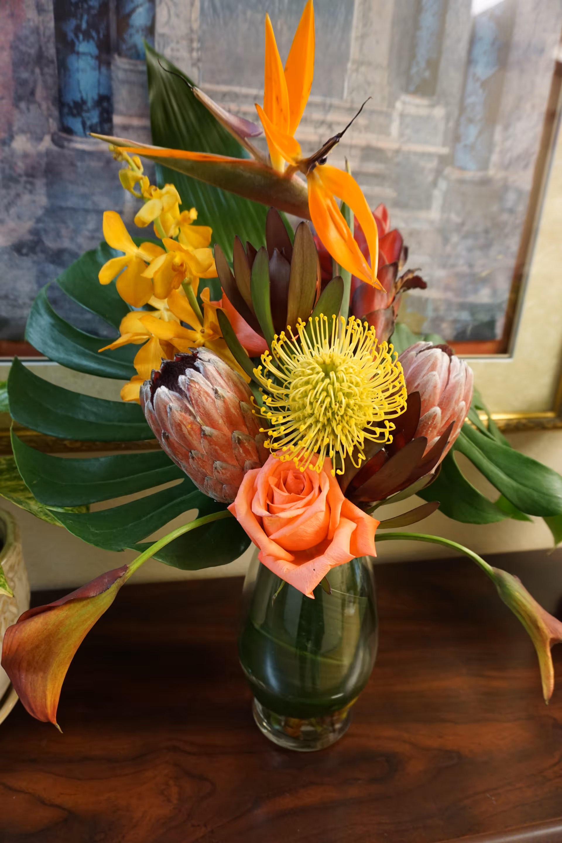 A vibrant floral arrangement in a glass vase on a wooden surface, featuring a variety of flowers including a bird of paradise, yellow orchids, protea, a yellow pincushion flower, an orange rose, and large green leaves, with a framed picture in the background.