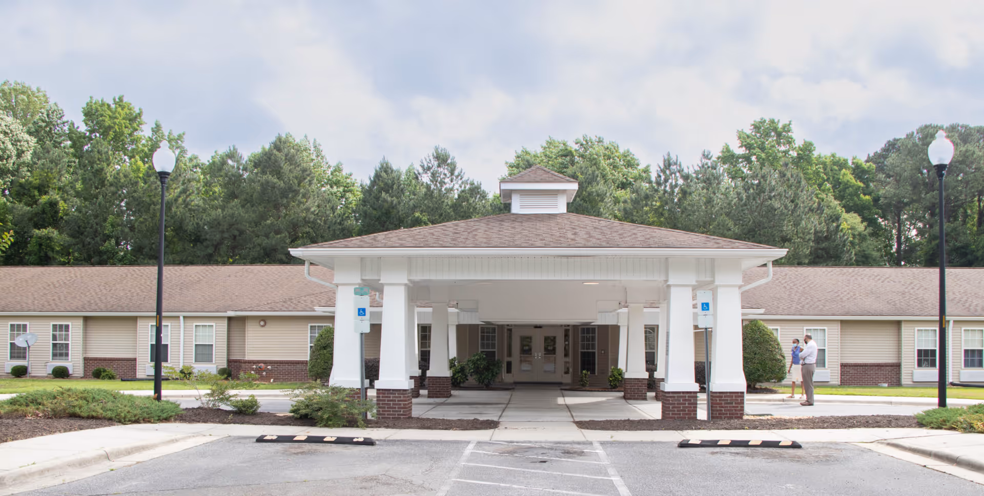 Covered entrance canopy of a single-story senior living building with a driveway, two lampposts, and trees in the background.