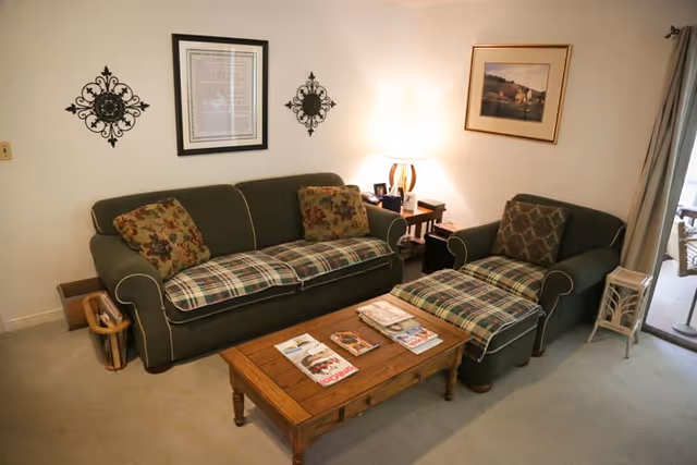 A cozy living room with a green sofa and matching armchair, both with plaid cushions. A wooden coffee table in front of the sofa holds several magazines. The walls are decorated with framed artwork and decorative metal wall hangings. A side table with a lamp and picture frames is next to the armchair. A sliding glass door with curtains is visible on the right side.