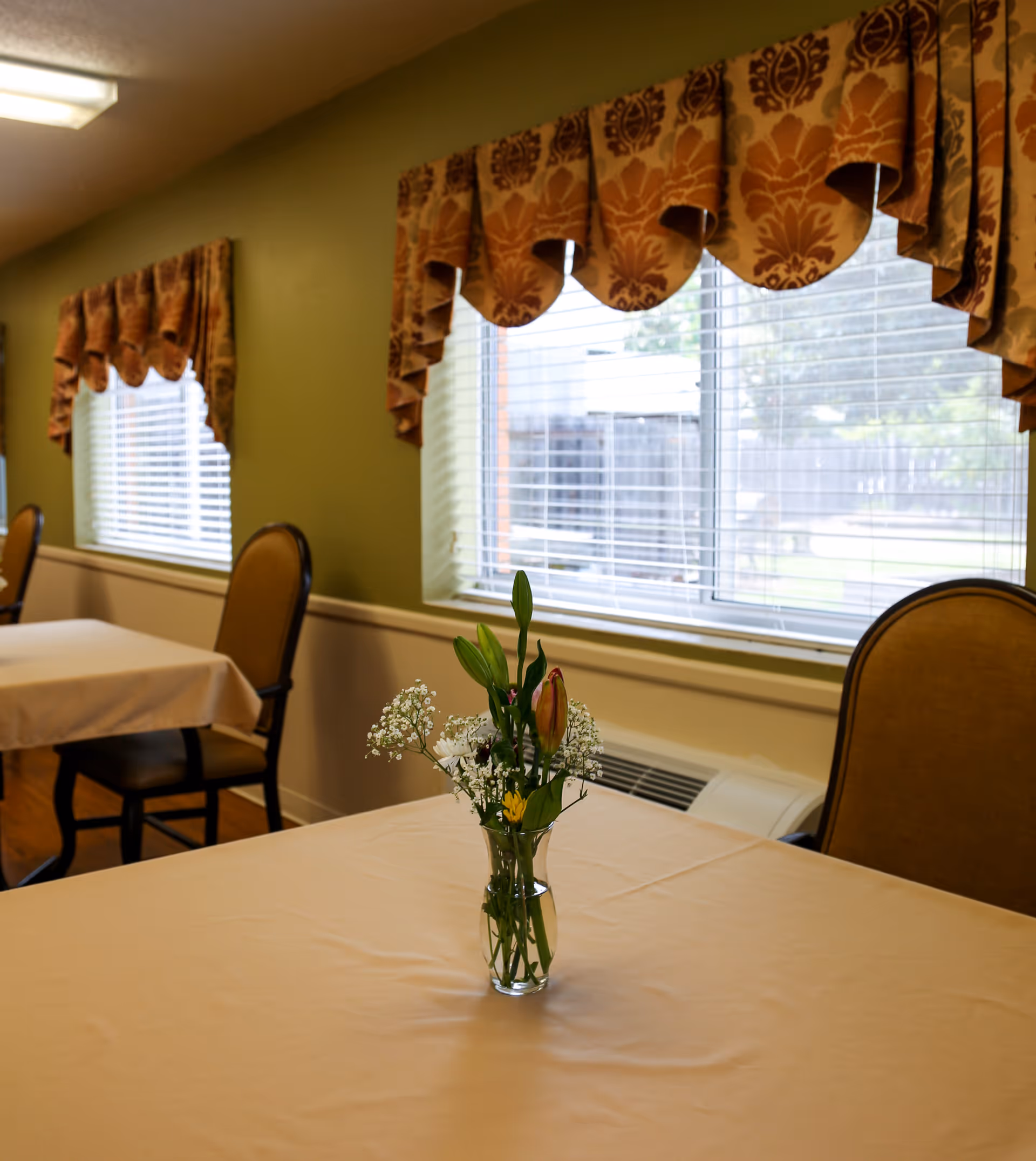 A dining area with tables covered in beige tablecloths, each table having a small vase with flowers. The room has green walls with two windows adorned with patterned valances, and chairs with cushioned backs and seats.