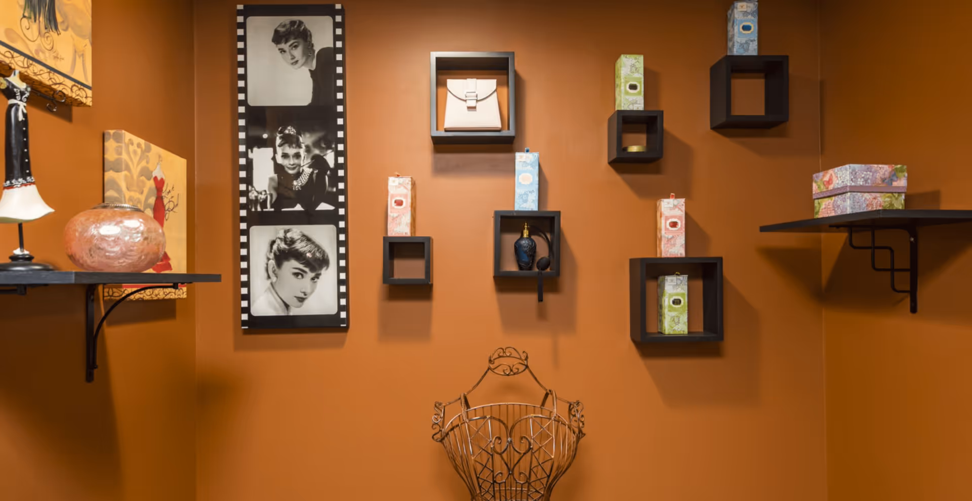 An interior wall painted in warm brown with decorative black square shelves holding various small items including boxes and a perfume bottle. A black and white photo collage of Audrey Hepburn is mounted on the wall. There are two black wall shelves on either side holding decorative items, and a decorative metal wire basket is positioned below the shelves.