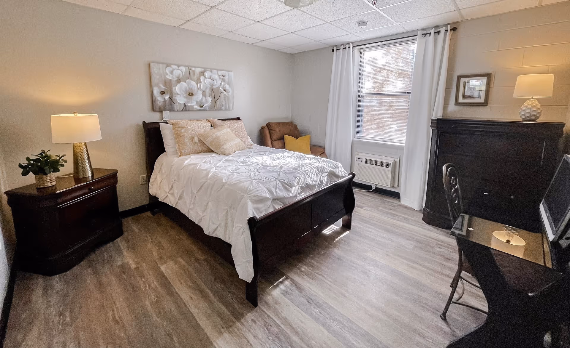 A cozy senior living bedroom with a wooden bed frame, white bedding, and decorative pillows. There is a nightstand with a lamp and a small plant on the left side of the bed. A window with white curtains lets in natural light, and an air conditioning unit is installed below the window. A brown armchair with a yellow pillow is placed in the corner. On the right side, there is a dark wooden dresser with a lamp and a framed picture above it, and a glass-top desk with a chair in front of it.