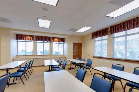 A bright room with multiple rectangular tables and blue chairs arranged in rows. The room has large windows with red patterned valances, beige walls, and a ceiling with recessed lighting panels.