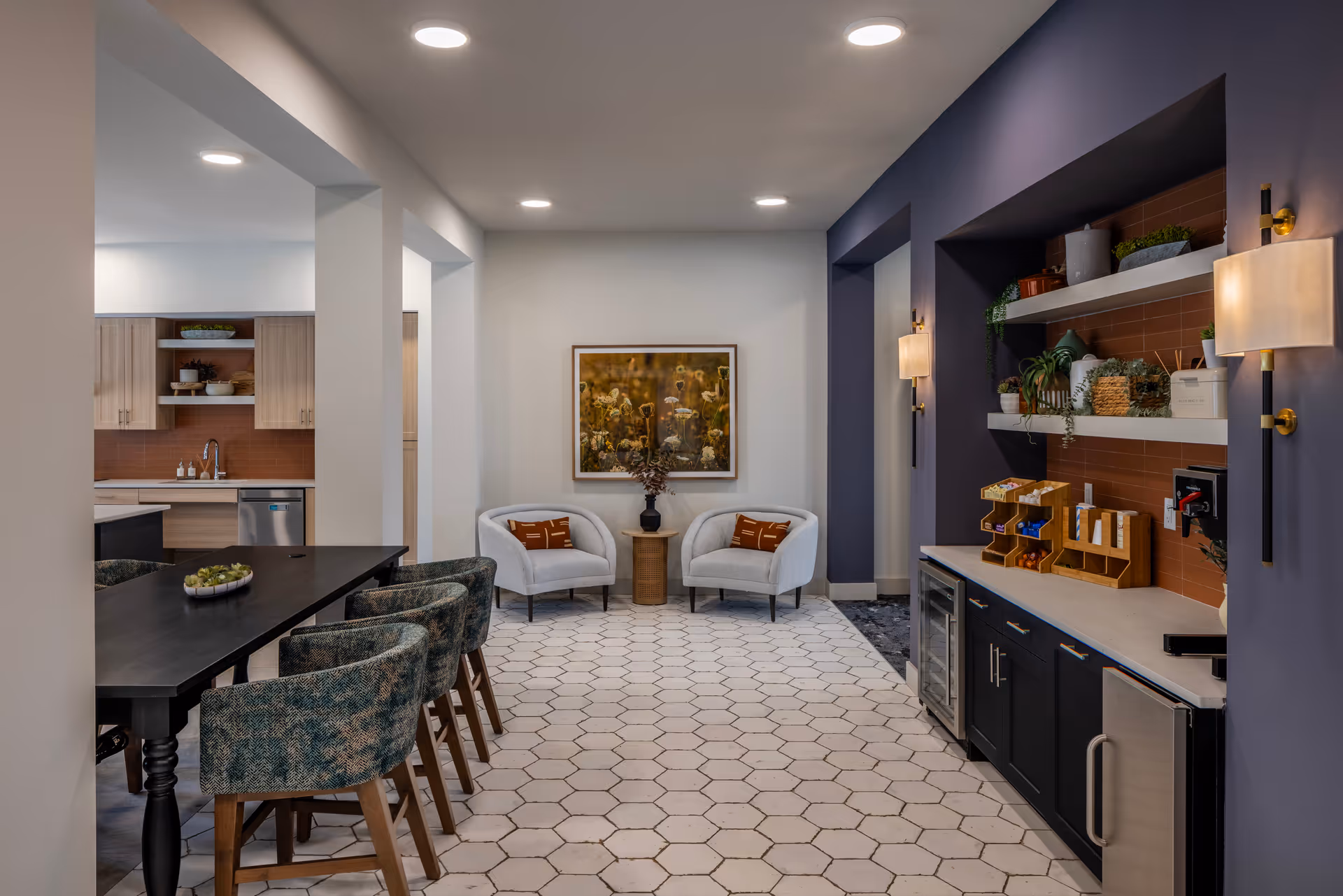 Interior view of a modern senior living facility common area featuring a black dining table with six upholstered chairs on the left, a small kitchen area with wooden cabinets and a stainless steel dishwasher in the background, and a cozy seating nook with two white armchairs and a small round table with a vase in the center. On the right, there is a counter with shelves holding plants and various items, under soft wall-mounted lights, and a hexagonal tiled floor throughout.