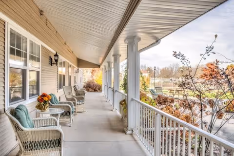 Covered outdoor porch with white wicker chairs and small tables arranged along the side of a building. The porch overlooks a garden area with trees, shrubs, and a picnic table under a partly cloudy sky.