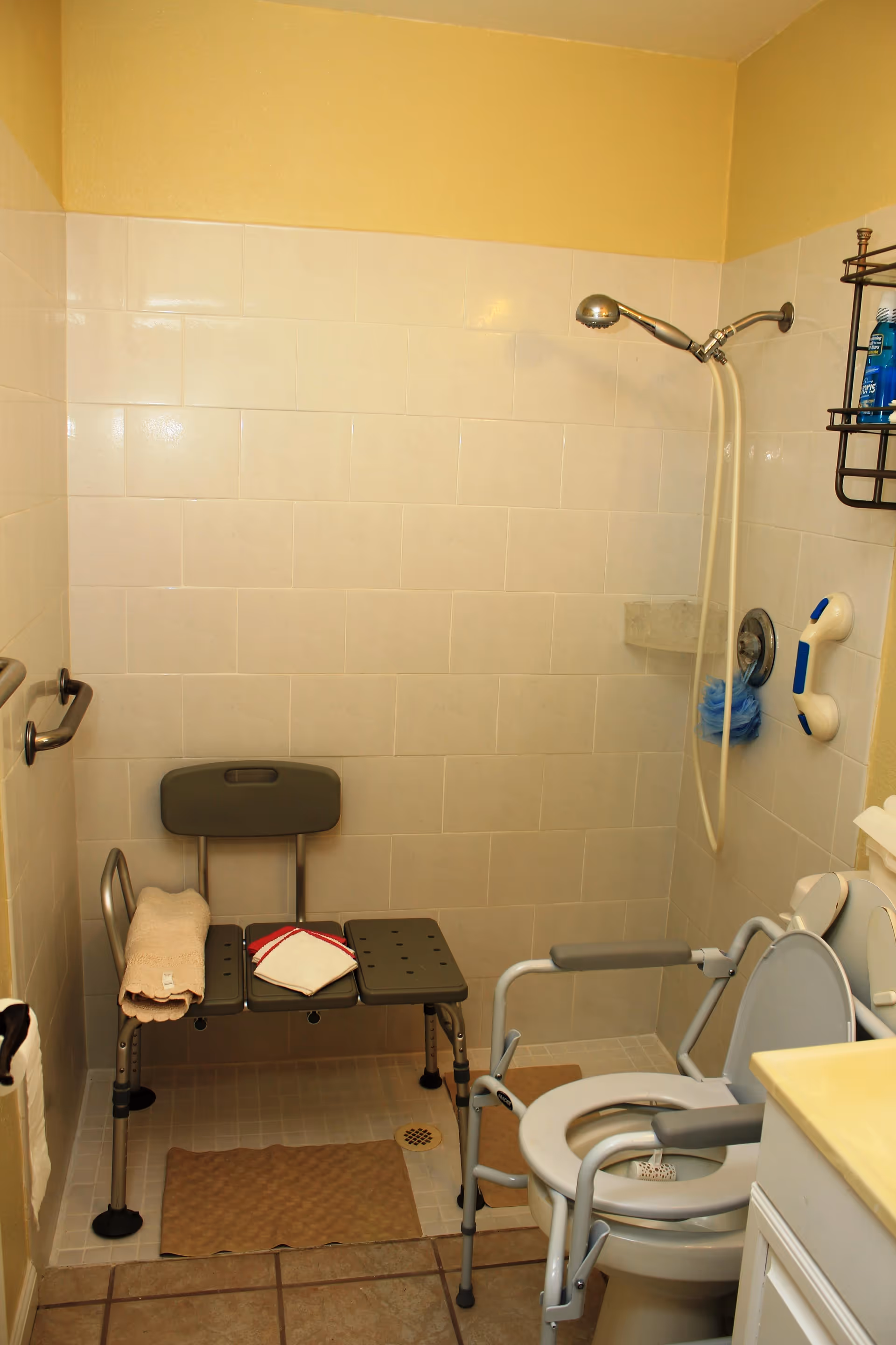 A bathroom with beige tiled walls and floor, featuring a shower area with a handheld showerhead and a shower bench with towels on it. There is a raised toilet seat with armrests, a grab bar on the wall, and a small shelf with toiletries.