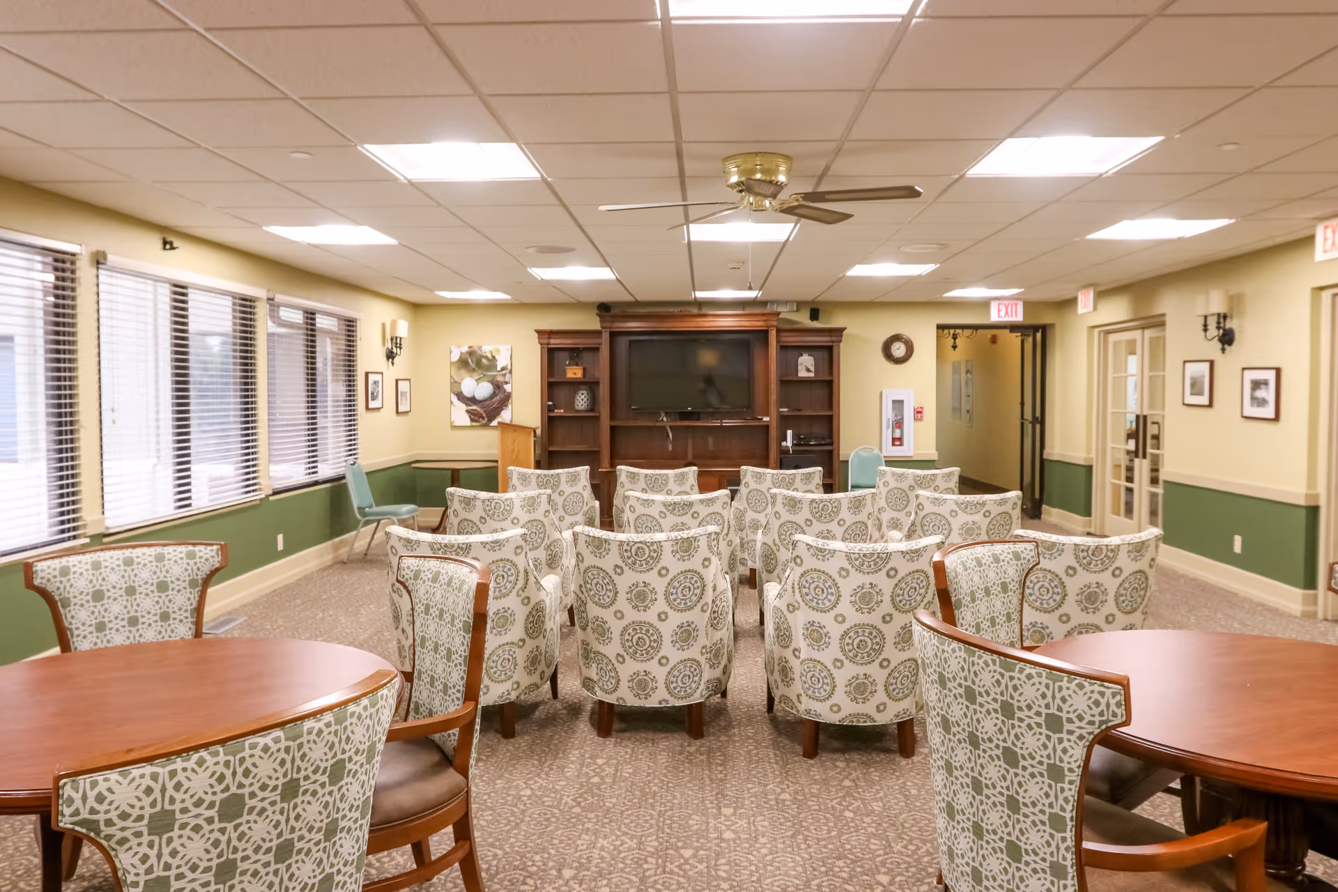 A well-lit common area with patterned armchairs arranged in rows facing a large wooden entertainment center with a flat-screen TV. The room has large windows with blinds on the left side, round tables with matching patterned chairs in the foreground, and beige walls with green wainscoting. Ceiling lights and a ceiling fan are visible above.