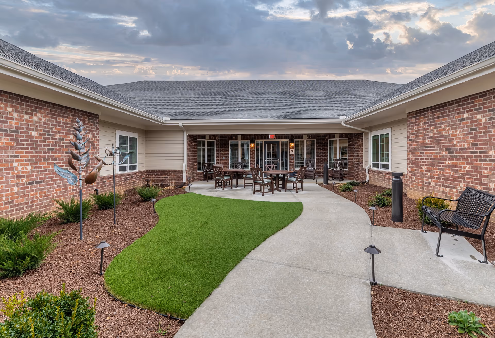 Outdoor courtyard area at The Capstone at Station Camp featuring a curved concrete walkway, green grass patch, brown mulch beds with plants and metal sculptures, several wooden tables and chairs on a concrete patio, and a black metal bench. The building has red brick walls with beige siding and multiple windows under a gray shingled roof, with a cloudy sky overhead.
