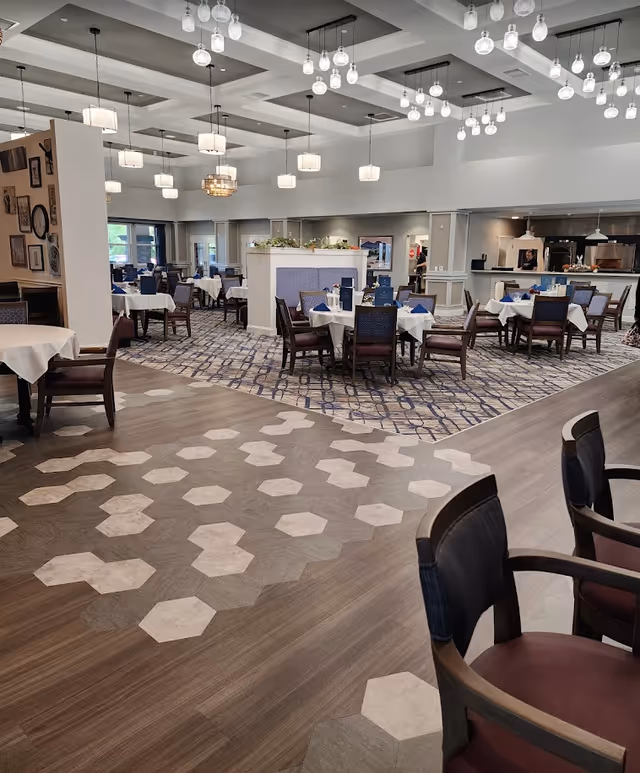 Spacious dining room in a senior living facility with multiple tables set with white linens, patterned flooring, and modern pendant lighting.