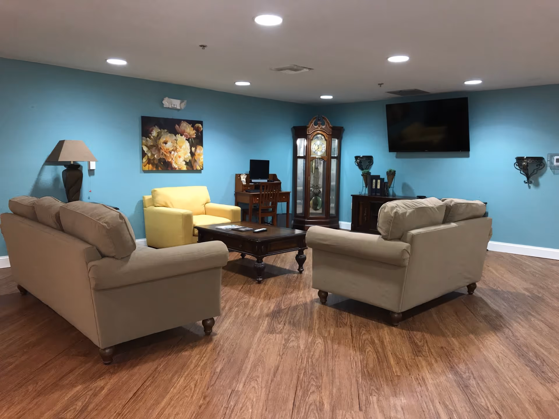 A senior living facility common area with two beige sofas and one yellow armchair arranged around a dark wooden coffee table on a wood floor. The walls are painted light blue and decorated with a floral painting, a tall grandfather clock, a wall-mounted TV, and a small wooden desk with a computer. There is a floor lamp on the left side and small decorative shelves on the right wall.