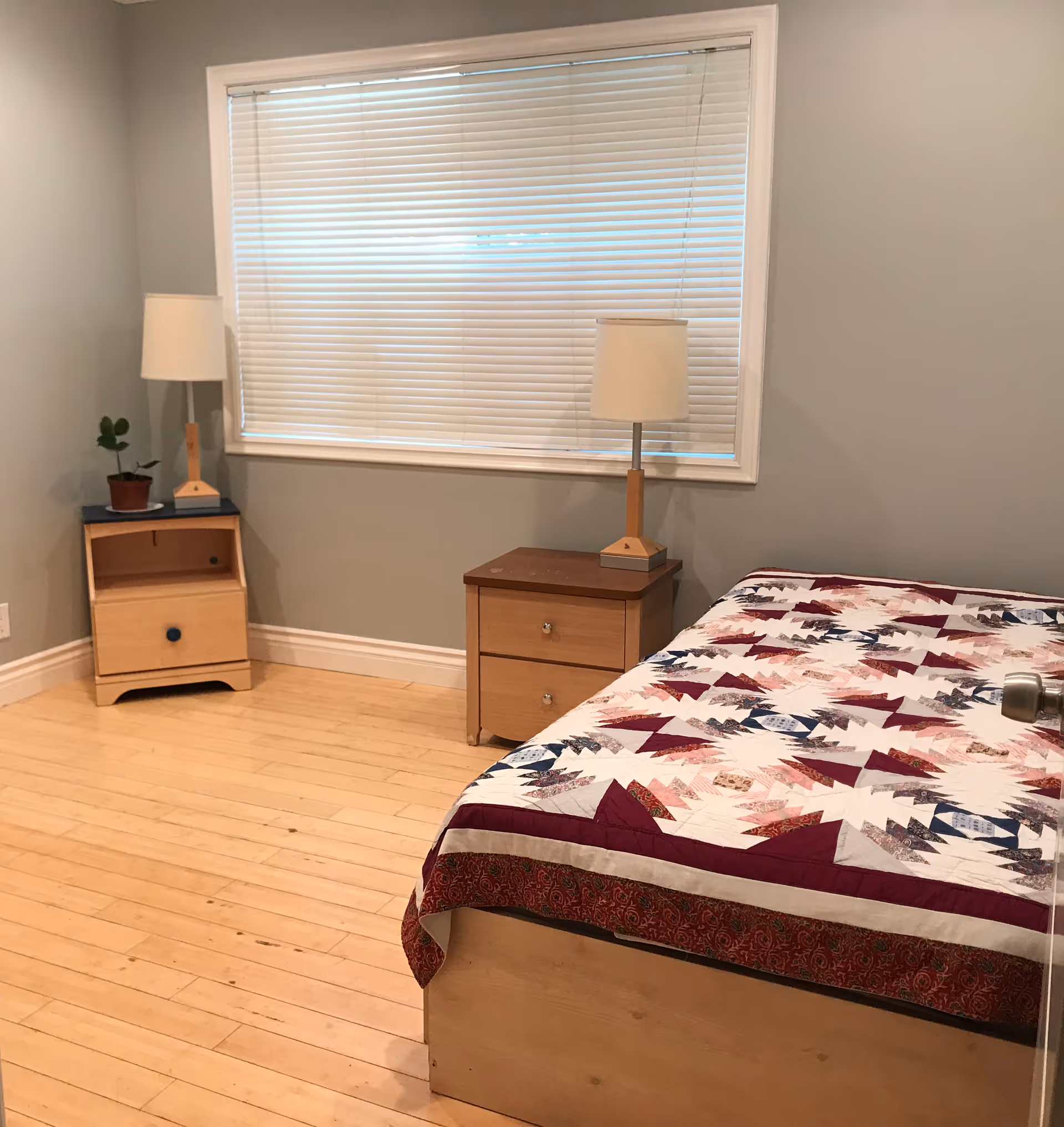 A simple bedroom with light wooden flooring, a bed with a colorful quilt, two wooden nightstands each with a lamp, a small potted plant on one nightstand, and a window with closed white blinds.