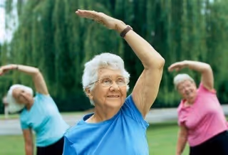 Three elderly women outdoors performing a stretching exercise with one arm raised above their heads, smiling and wearing casual clothing in blue, teal, and pink shirts, with greenery in the background.