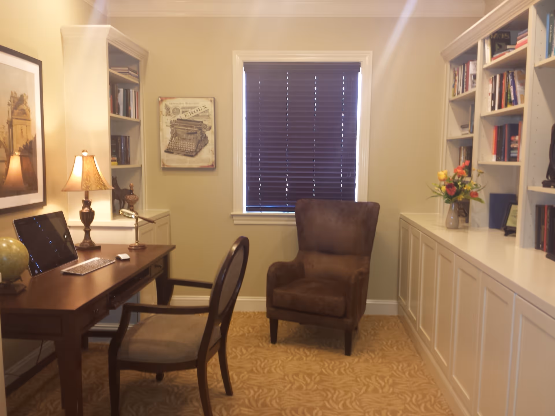 Small office/reading room with a wooden desk and chair, an upholstered armchair by a window with closed blinds, and built-in bookshelves.