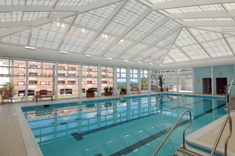 Indoor swimming pool area with clear blue water, surrounded by large windows letting in natural light. There are benches and potted plants along the perimeter, and a high, white, vaulted ceiling with grid-like panels. The pool has metal handrails and steps for easy access.