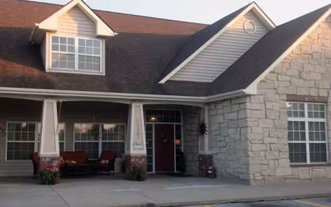 Exterior view of a senior living facility entrance with porch seating and stone walls.