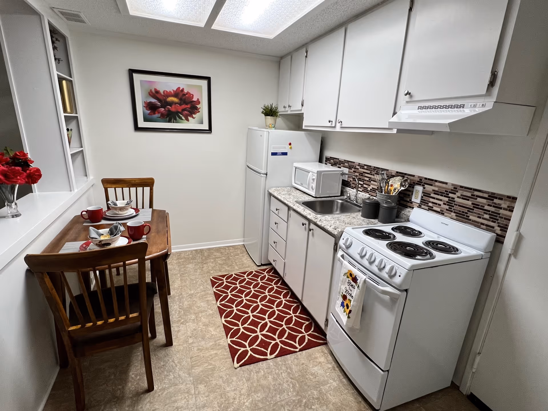 A small kitchen area with white cabinets, a white refrigerator, microwave, and electric stove. There is a red patterned rug on the floor in front of the sink and stove. A wooden dining table with two chairs is set with red mugs, bowls, and napkins. A framed picture of a red flower hangs on the wall above the refrigerator. The kitchen has a tiled backsplash and beige flooring.