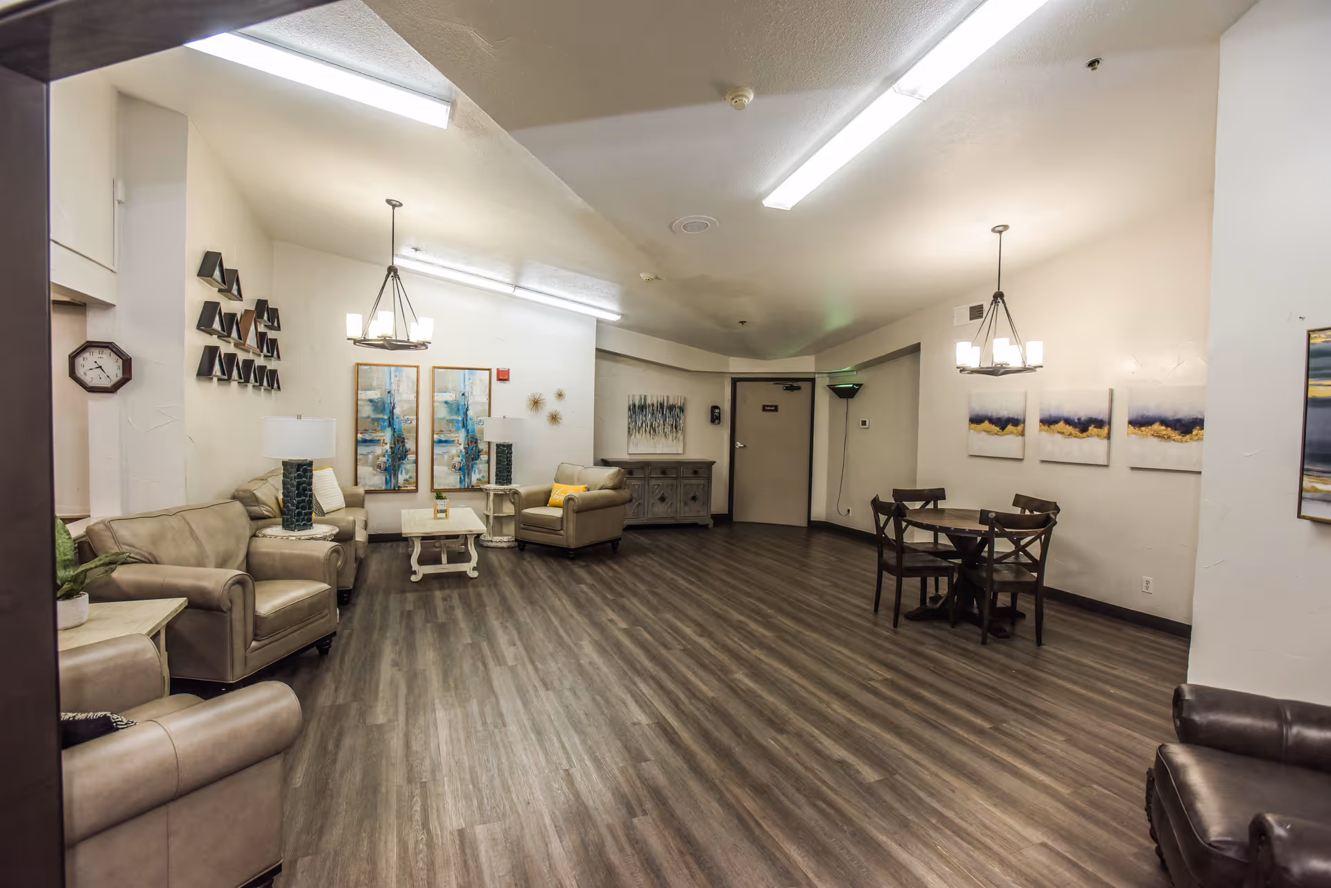 A spacious common area in Graham Oaks Care Center featuring beige leather sofas and armchairs arranged around a white coffee table on the left side. On the right side, there is a small wooden dining table with four chairs. The room has wood-look flooring, white walls adorned with modern artwork, and two ceiling light fixtures. A clock is mounted on the left wall near an open doorway.