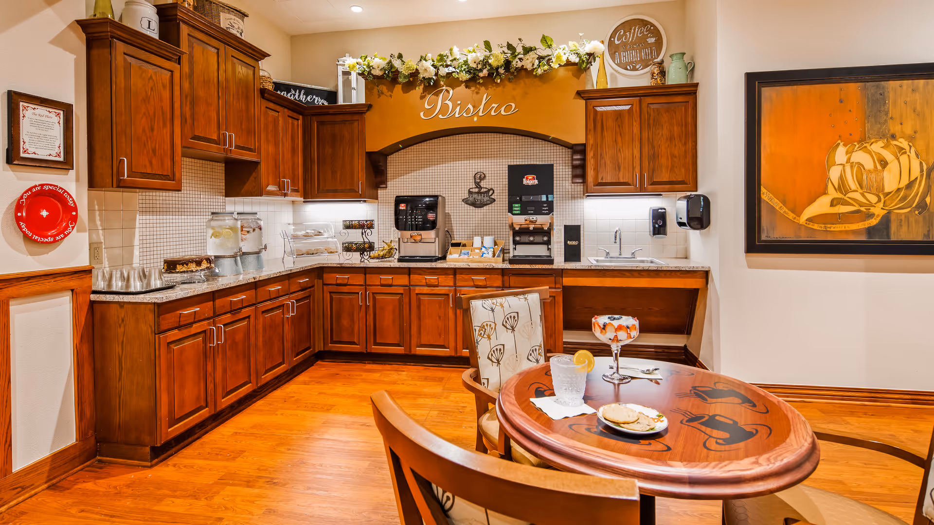 A cozy bistro area with wooden cabinets and countertops, featuring a coffee machine, water dispensers, and a small sink. A round wooden table with two chairs is set with a glass of water with lemon, a dessert dish, and a small plate with a pastry. The wall above the counter has a decorative sign that says 'Bistro' with a floral garland. There is a framed artwork on the right wall and a red decorative plate on the left wall.