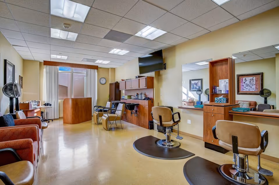 Interior view of a salon area in a senior living facility with multiple styling chairs, mirrors, hair drying stations, and a television mounted on the wall. The room has beige walls, a tiled ceiling with fluorescent lighting, and a reception desk near a window with vertical blinds.