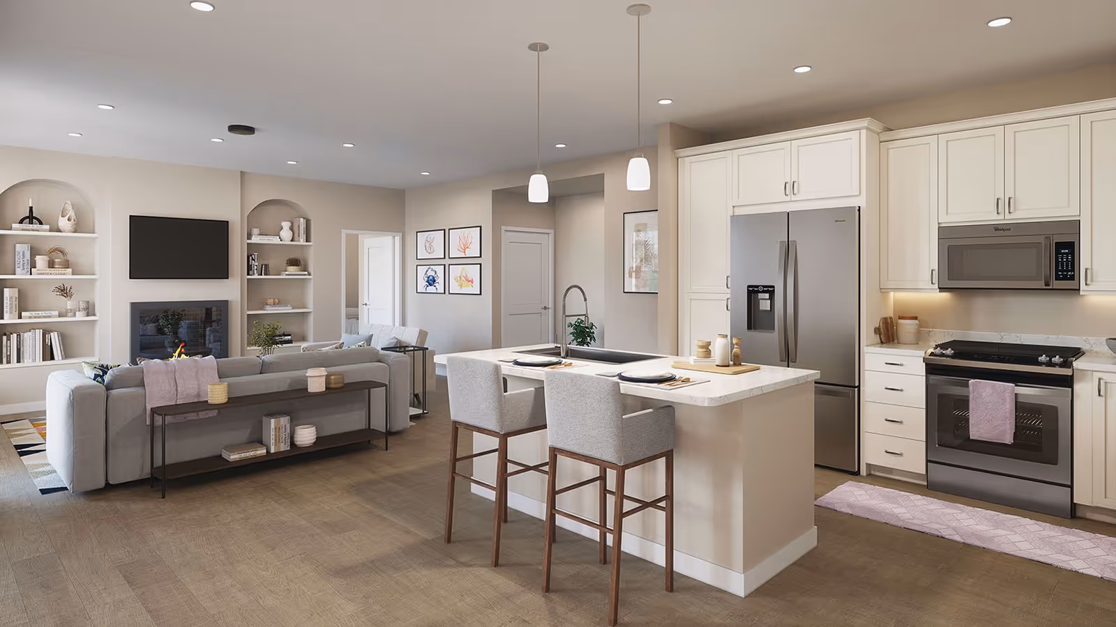 A modern open-concept living area and kitchen in a senior living community. The kitchen features white cabinetry, stainless steel appliances including a refrigerator, microwave, and oven, and a large island with a sink and two upholstered bar stools. The living area has a light gray sectional sofa facing a wall-mounted TV above a fireplace, with built-in shelves on either side. The space is decorated with framed artwork, plants, and soft lighting from recessed ceiling lights and pendant lamps over the island.