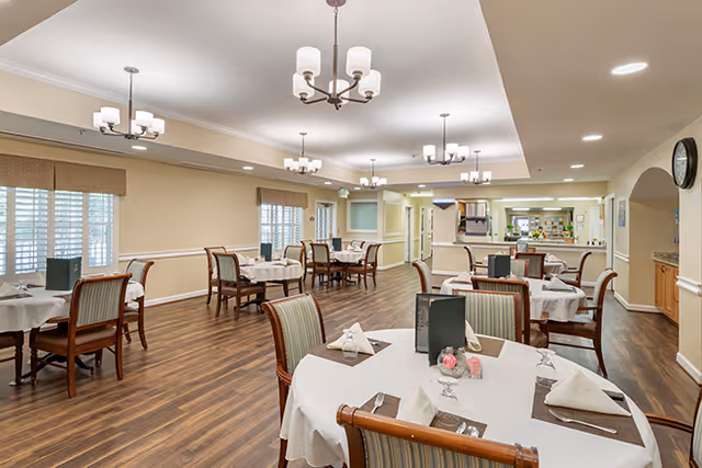 A spacious dining room with multiple round tables covered with white tablecloths, each set with napkins, glasses, and menus. The room has wooden flooring, beige walls, and several chandeliers hanging from the ceiling. Large windows with blinds allow natural light to enter the room.