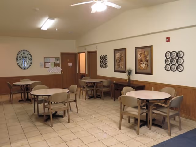A dining room with several round tables and beige chairs arranged around them. The room has tiled floors, wood paneling on the lower half of the walls, and light-colored upper walls. There are framed pictures and decorative wall art on the walls, a large clock, and a bulletin board. Ceiling lights and a ceiling fan illuminate the space.