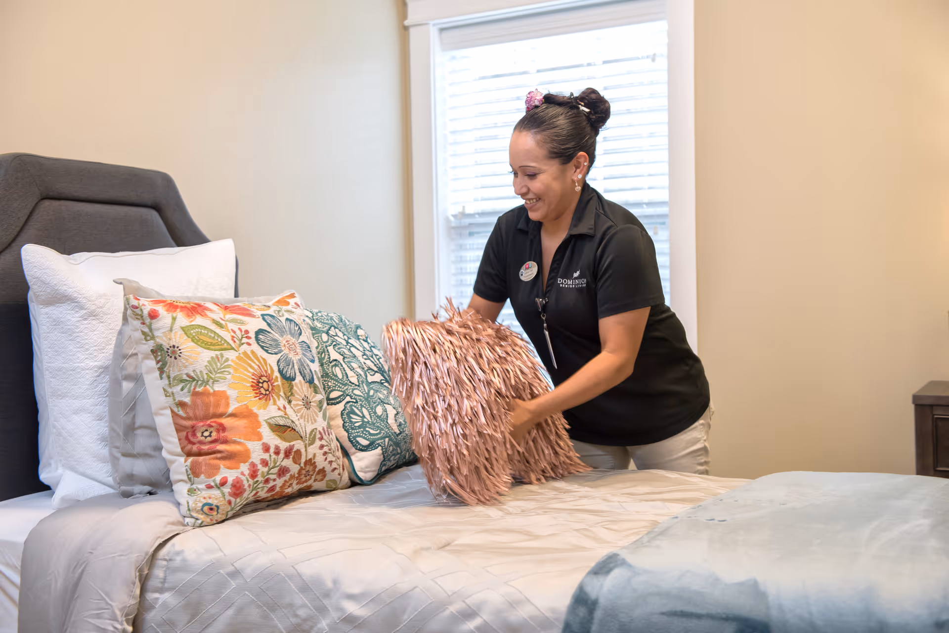 A woman wearing a black Dominion Senior Living polo shirt is smiling while arranging decorative pillows on a neatly made bed in a bedroom with beige walls and a window with white blinds.